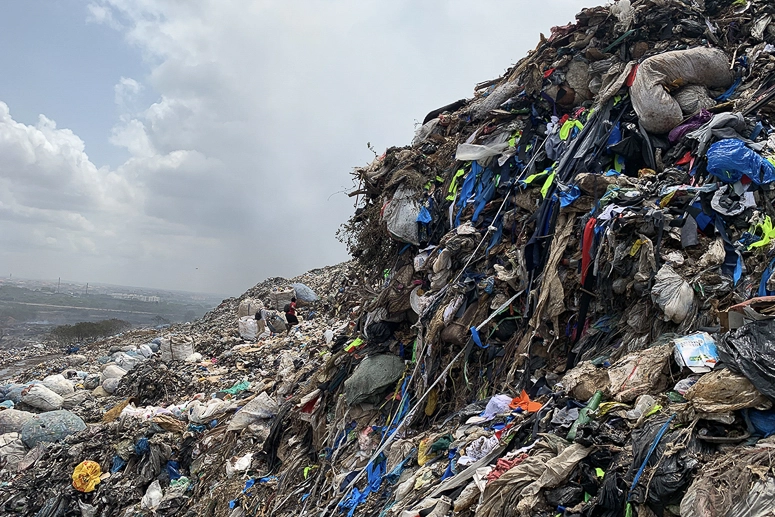 En gigantisk losseplads fyldt med tekstilaffald og brugt tøj i Ghana. Farvede tekstiler, plastik og affald ligger i store bunker med mennesker i baggrunden.